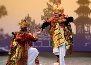 Two actors dressed in royal Balinese attire in front of a shadow landscape of trees and buildings.