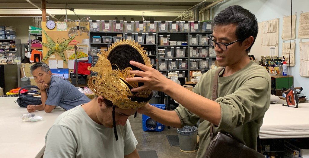 A master artist fits a Balinese headdress on a performer with another artist observing.