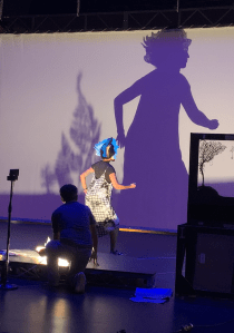 A performer wearing a 3D wayang listrik mask casts a shadow on a screen with a teacher observing in the background.